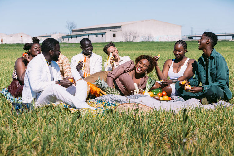 Ground level of group of stylish black woman with banana smiling with closed eyes and raising arm while lying on blanket amidst friends during picnic in grassy field on sunny summer day