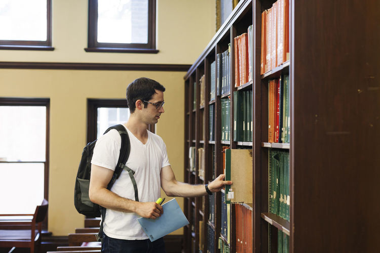 Confident male student with backpack searching for books in shelf at library