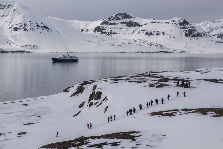 Group of explorers walking on snowcapped shoreline of arctic ocean with ship in the background.