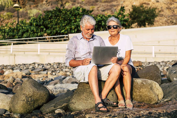 Full length of couple using laptop while sitting on rock