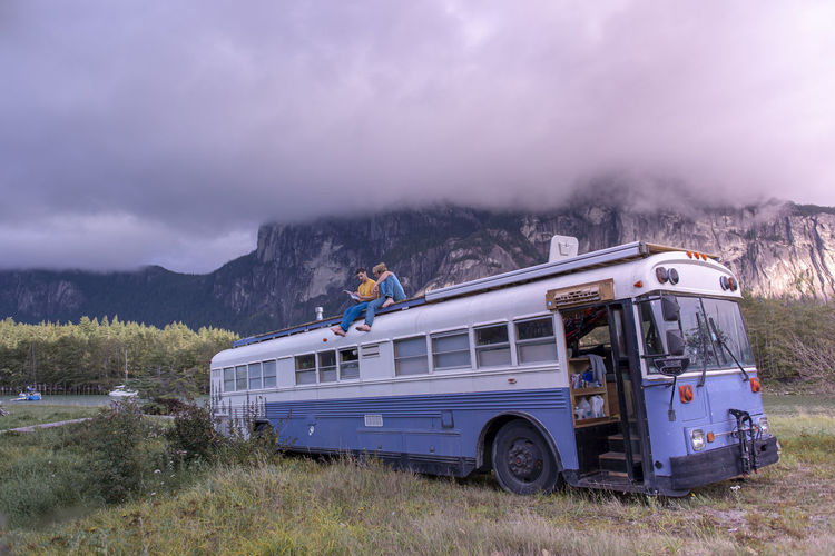 Two people sitting on top of school bus reading book front of mountain