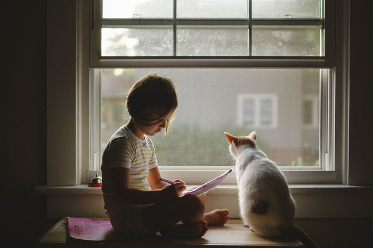 A little girl and her cat sit together peacefully on a window bench