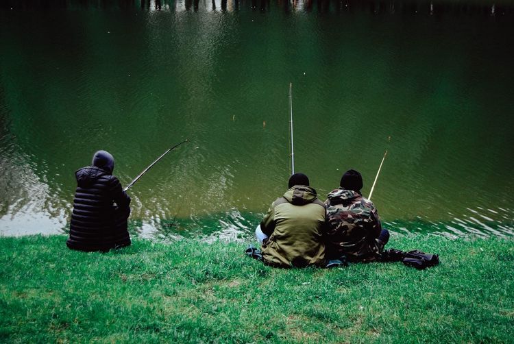 Rear view of men sitting on fishing rod by lake