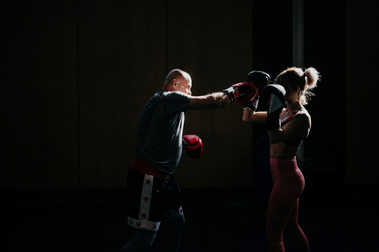 Side view of concentrated young sportswoman athlete in boxing gloves training fighting with professional male coach in modern gym
