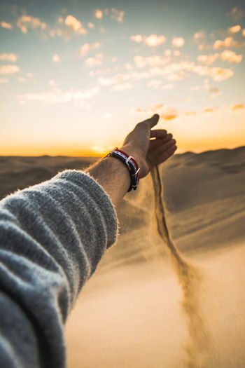 Cropped hand of man falling sand on desert against sky during sunset