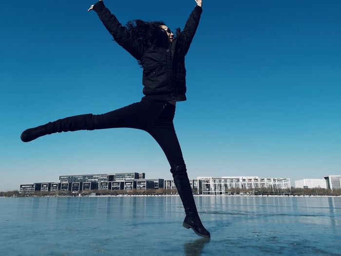 Woman jumping on frozen lake against clear blue sky
