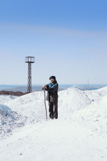 Man in ethnic clothes standing among white mounds and observation tower on mountain under blue sky.