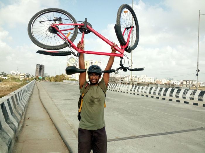 Happy cyclist carrying bicycle while standing on highway in city against sky