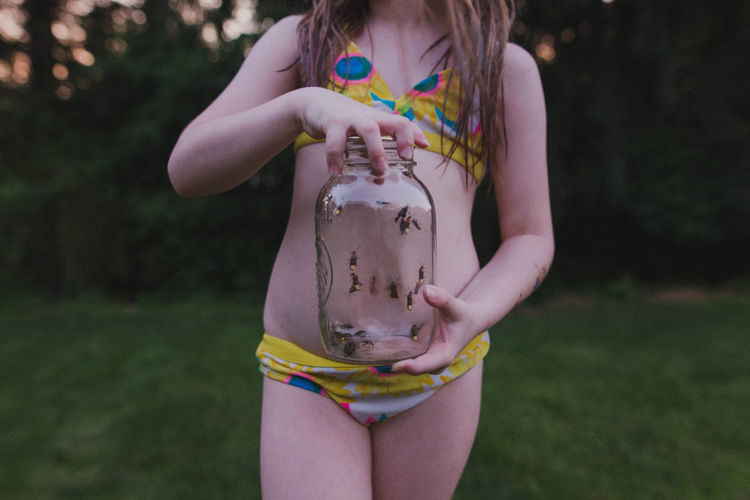Midsection of girl wearing bikini holding insects in jar while standing against trees at park