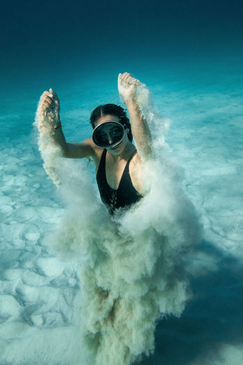 High angle of slim female in goggles and swimsuit swimming underwater and playing with sand while looking at camera