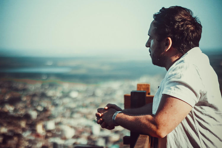 Side view of young man looking at cityscape against sky