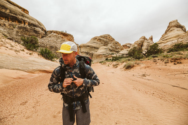 Man in hiking hat and camo jacket buckles his chest strap in desert