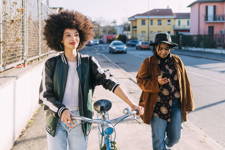 Young woman walking with bicycle by female friend on footpath