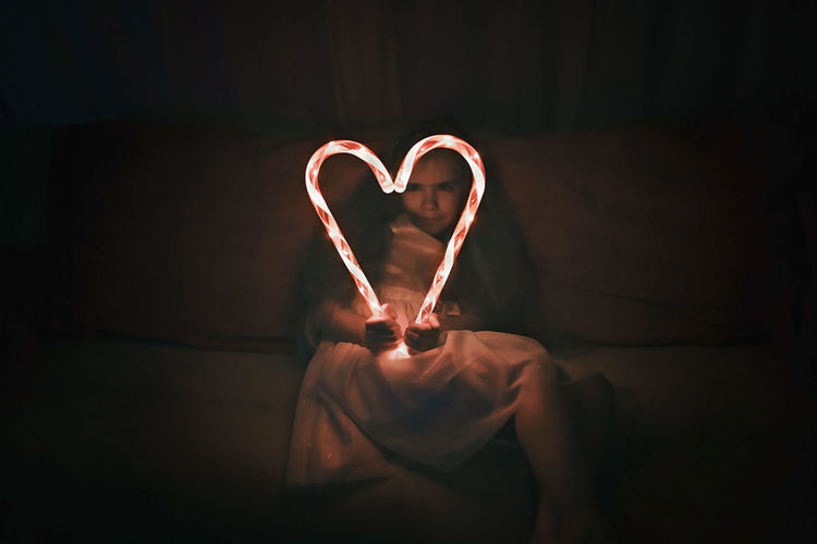 Girl making heart shape of illuminated candy canes on sofa in darkroom