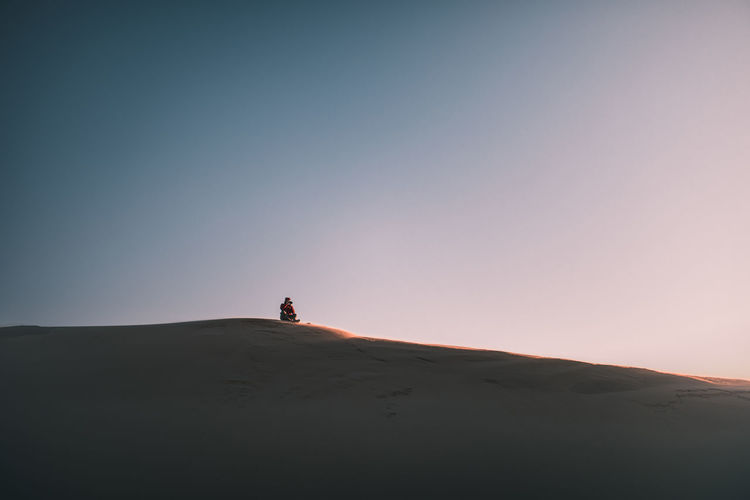 Distant view of man sitting on sand dune against clear sky