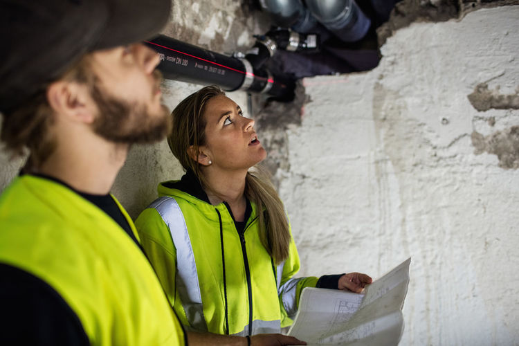 Concentrated male and female plumbers looking up while reading document at basement
