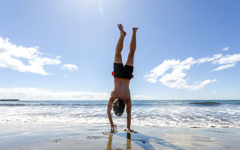 Rear view of shirtless man performing handstand at beach against sky