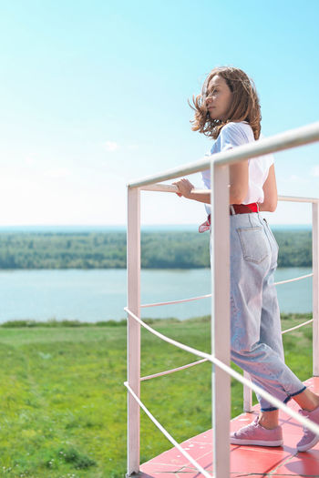 Portrait of young woman standing by railing against sky