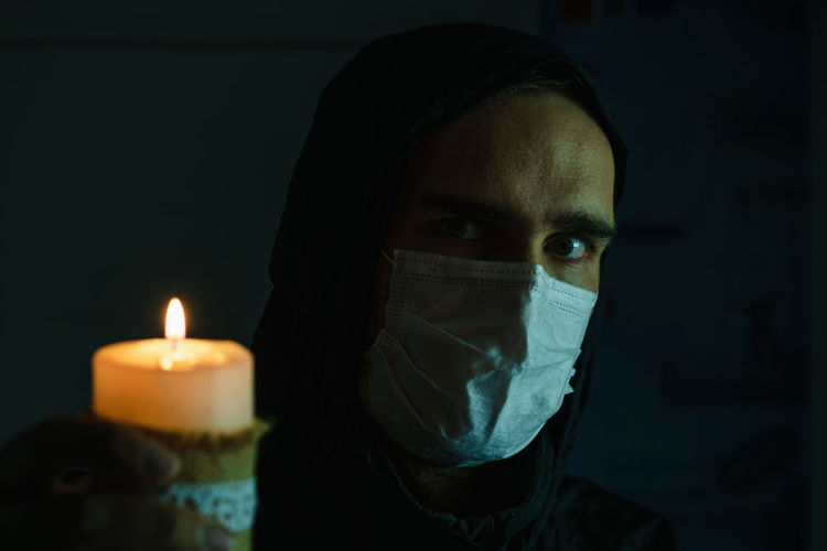 The face of a man wearing a medical mask and black hood against the background of a lit candle.