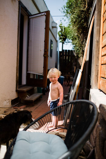 Shirtless two year old standing on side of house smiling at dog