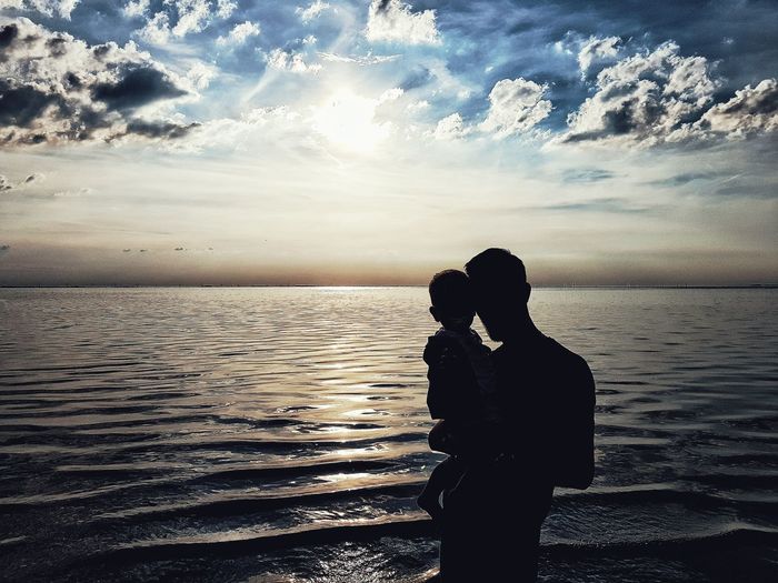 Rear view of silhouette couple standing at beach against sky during sunset