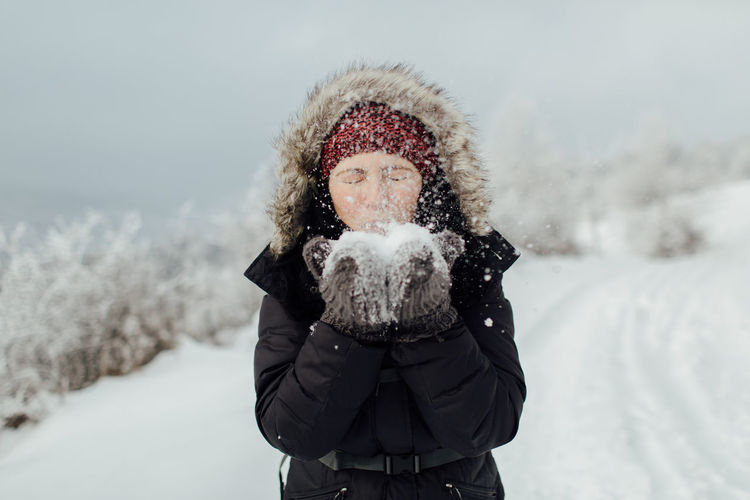 Playful woman blowing snow from hands while standing on mountain