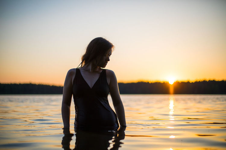 Side view of young woman standing in sea against sky during sunset