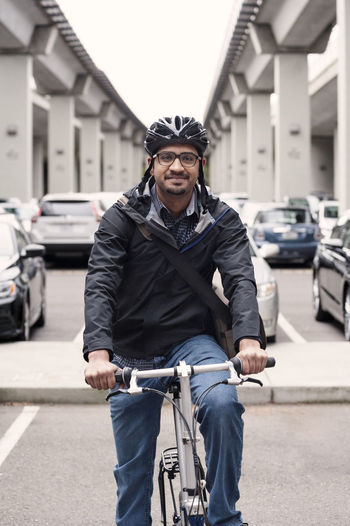 Portrait of smiling businessman sitting on bicycle with bridges and parking lot in background