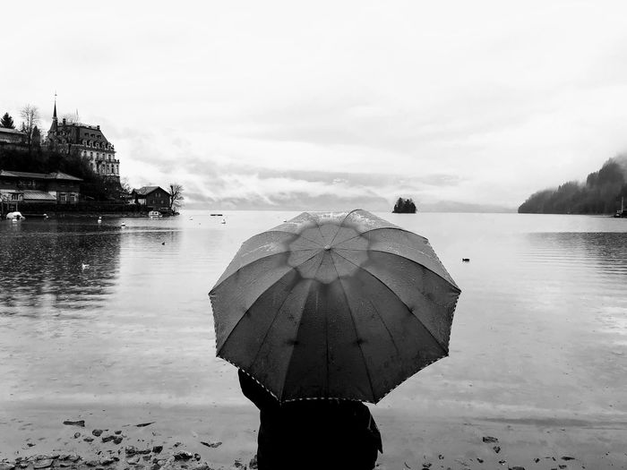 Rear view of person standing with umbrella at lakeshore against sky