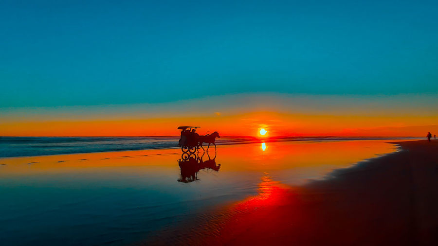 A silhouetted horse-drawn carriage at sunset time on the wet reflecting sand beach at parangtritis.