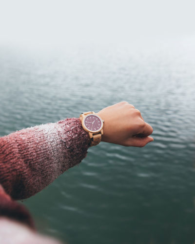 From above crop anonymous female in warm sweater checking time on stylish wristwatch while holding hand over river in cloudy day