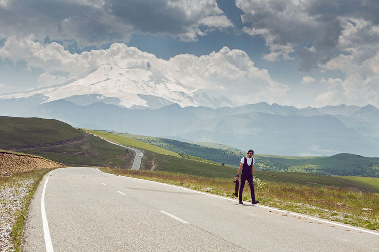 Guy in black jeans and a cap carries a skate on of mount everest in the summer on an asphalt road