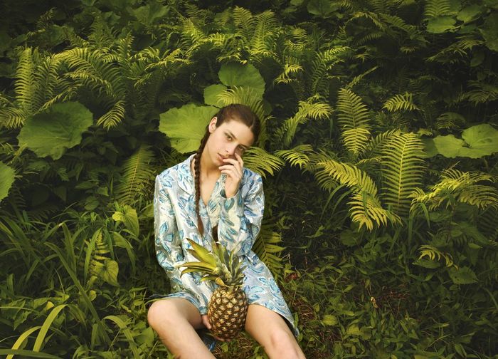 Portrait of young woman holding pineapple while sitting against plants