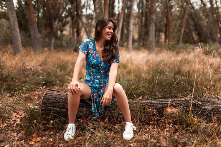 Full length of smiling young woman sitting on log in forest
