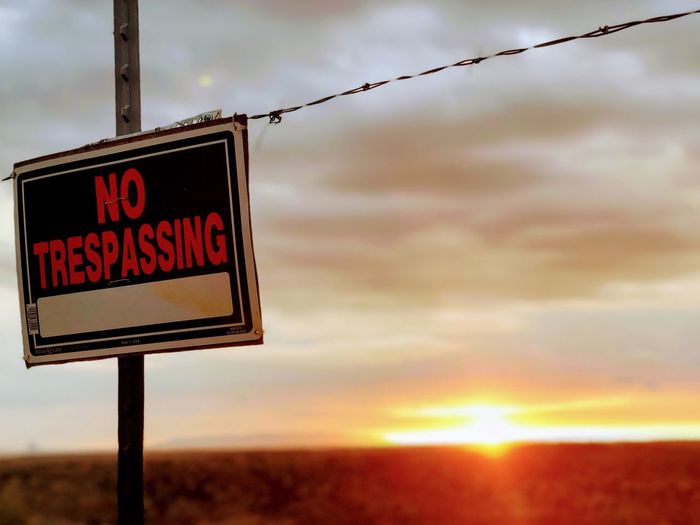 No trespassing sign on barbed wire against sky during sunset