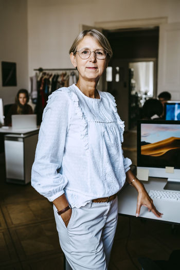 Mature businesswoman with hand in pocket standing by desk in office
