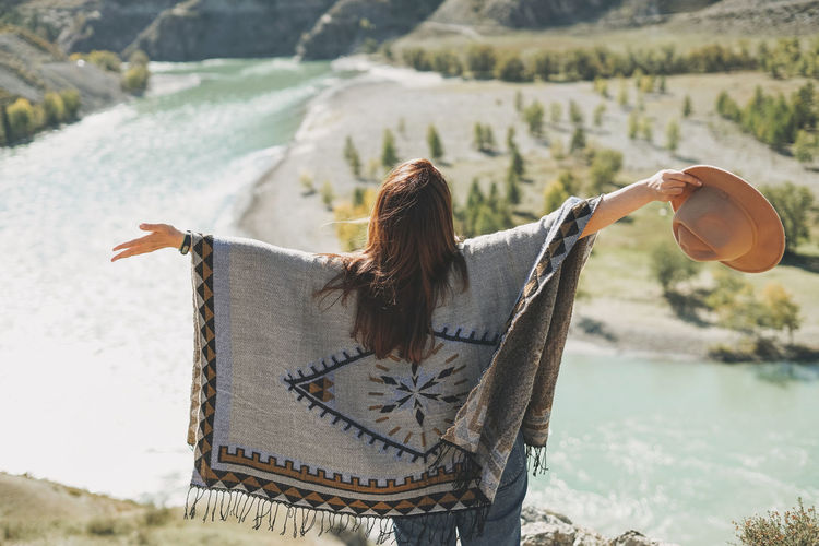 Carefree brunette young woman traveler in poncho from back on magic background of mountain river