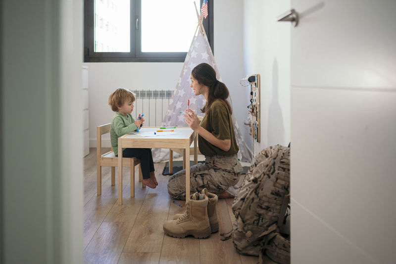 Side view of caring military mother in uniform and son drawing with colorful felt pens on paper in light room with camouflage backpack