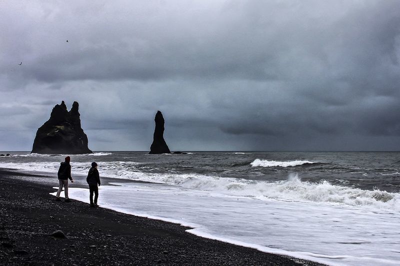 Male friends on shore against cloudy sky at vik