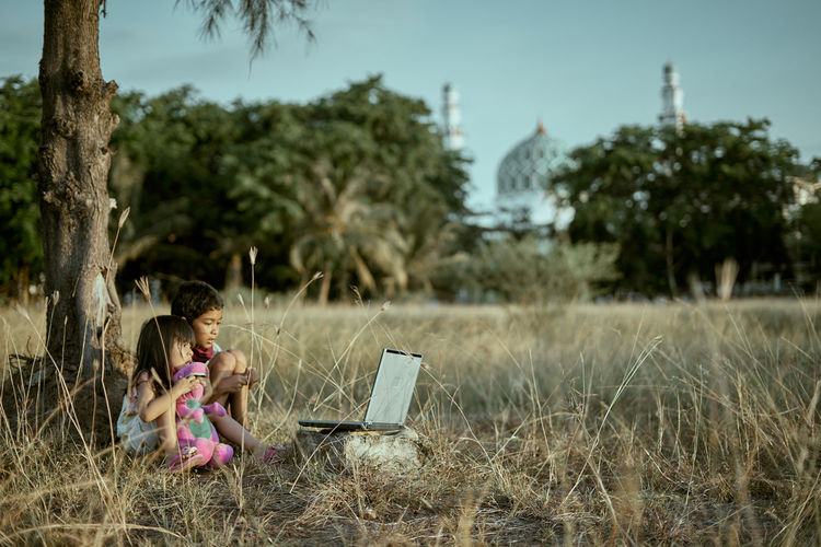 Siblings with laptop sitting on grassy land