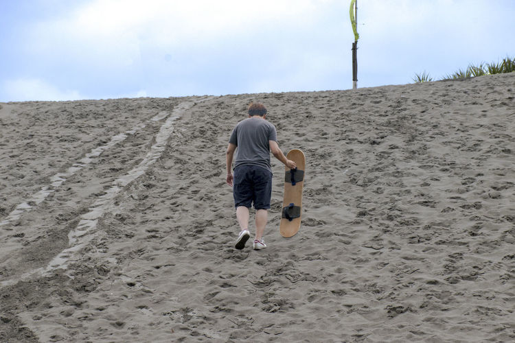 Rear view of man carrying sandboard on sandy hill against sky