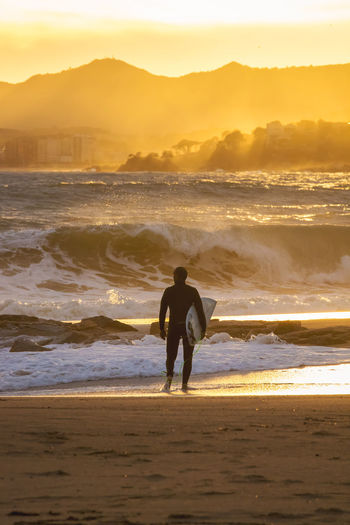 Rear view of man with surfboard standing on beach against sky during sunset
