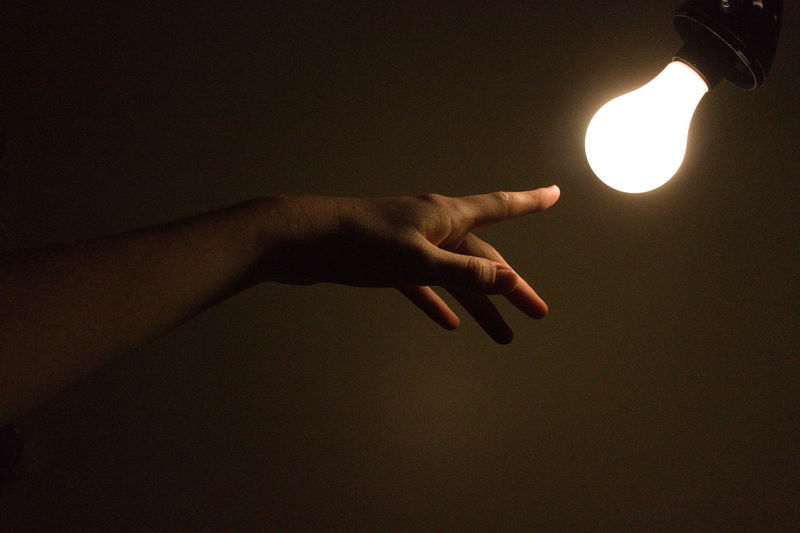 Cropped hand of person gesturing at illuminated light bulb against wall