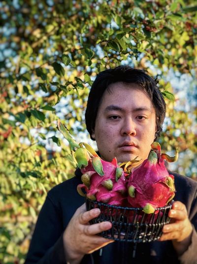 Portrait of young asian man holding basket of dragon fruits against trees.