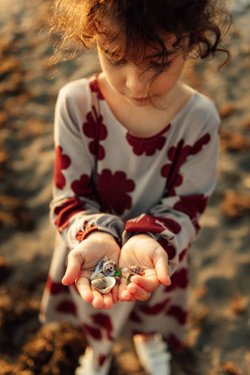 From above of little curly haired girl in dress demonstrating handful of seashells while spending summer day on beach