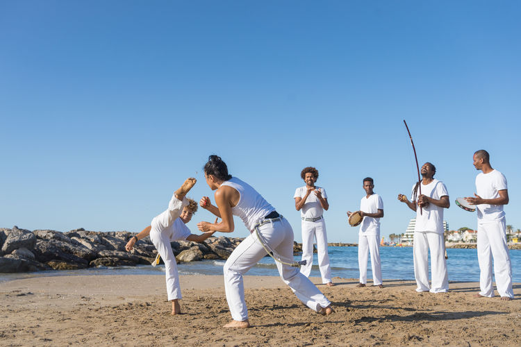 Group of anonymous barefoot black partners in white clothes practicing martial art on sandy beach under blue sky