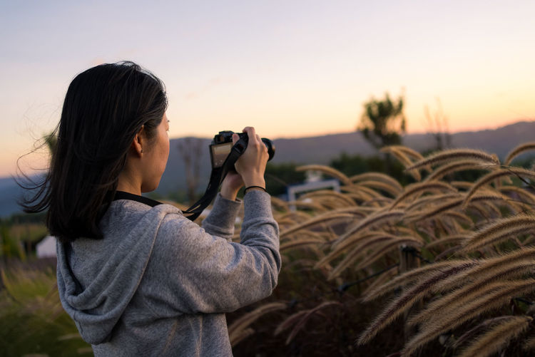 Side view of woman photographing against sky during sunset