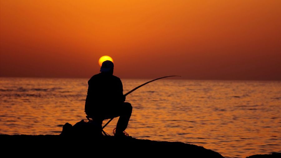 Silhouette man fishing in calm sea at sunset