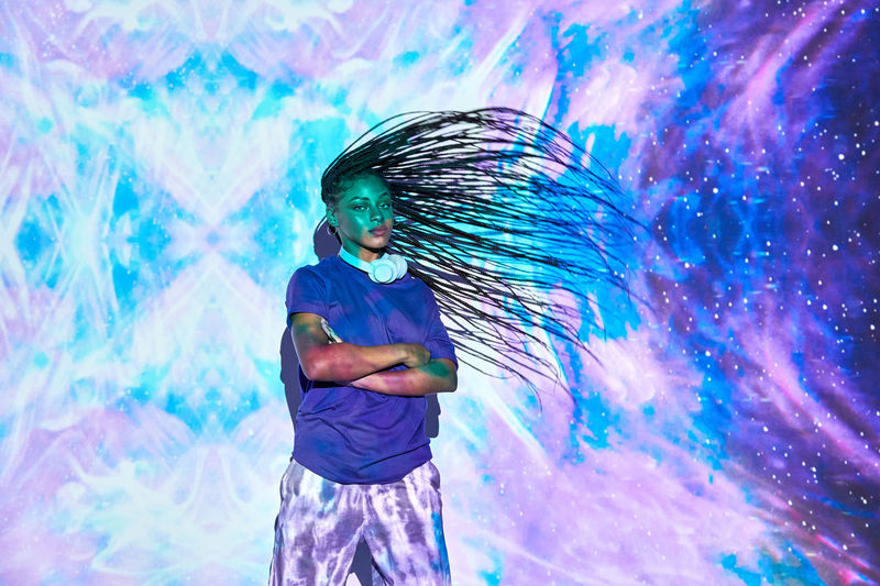 Side view of ethnic female millennial shaking long braided hair while his arms are crossed near wall with bright blue and lilac illumination