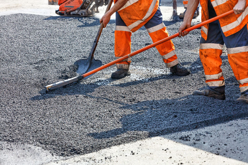 A working group of workers in orange overalls is repairing section of the road with fresh asphalt  